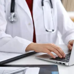 Lekarz w białym fartuchu z fonendoskopem pracuje przy laptopie i dokumentach.