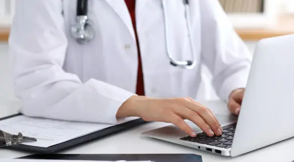 Lekarz w białym fartuchu z fonendoskopem pracuje przy laptopie i dokumentach.