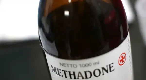 Butelka z ciemnobrązowym płynem, na etykiecie napis "Methadone" i czerwony symbol krzyża.