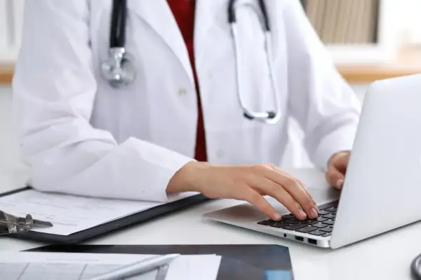 Lekarz w białym fartuchu z fonendoskopem pracuje przy laptopie i dokumentach.